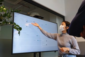Woman wearing face mask pointing at a large smart screen as part of her PowerPoint presentation.